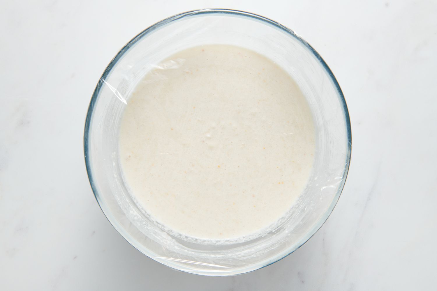 A large glass bowl of Sona Masuri rice-urad dal batter, covered with plastic wrap