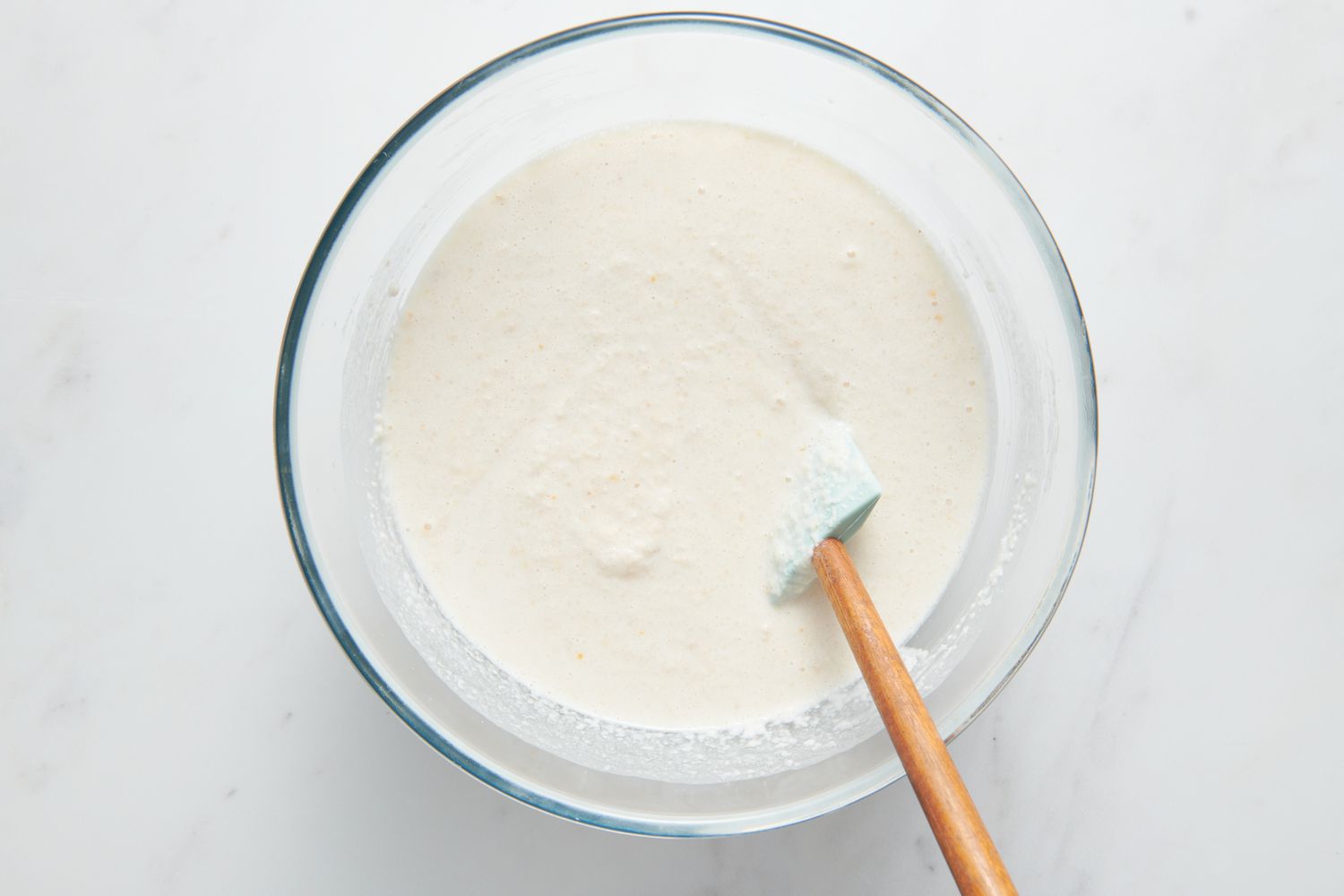 A large glass bowl with a silicone spatula folding the Sona Masuri rice batter into the urad dal-fenugreek batter