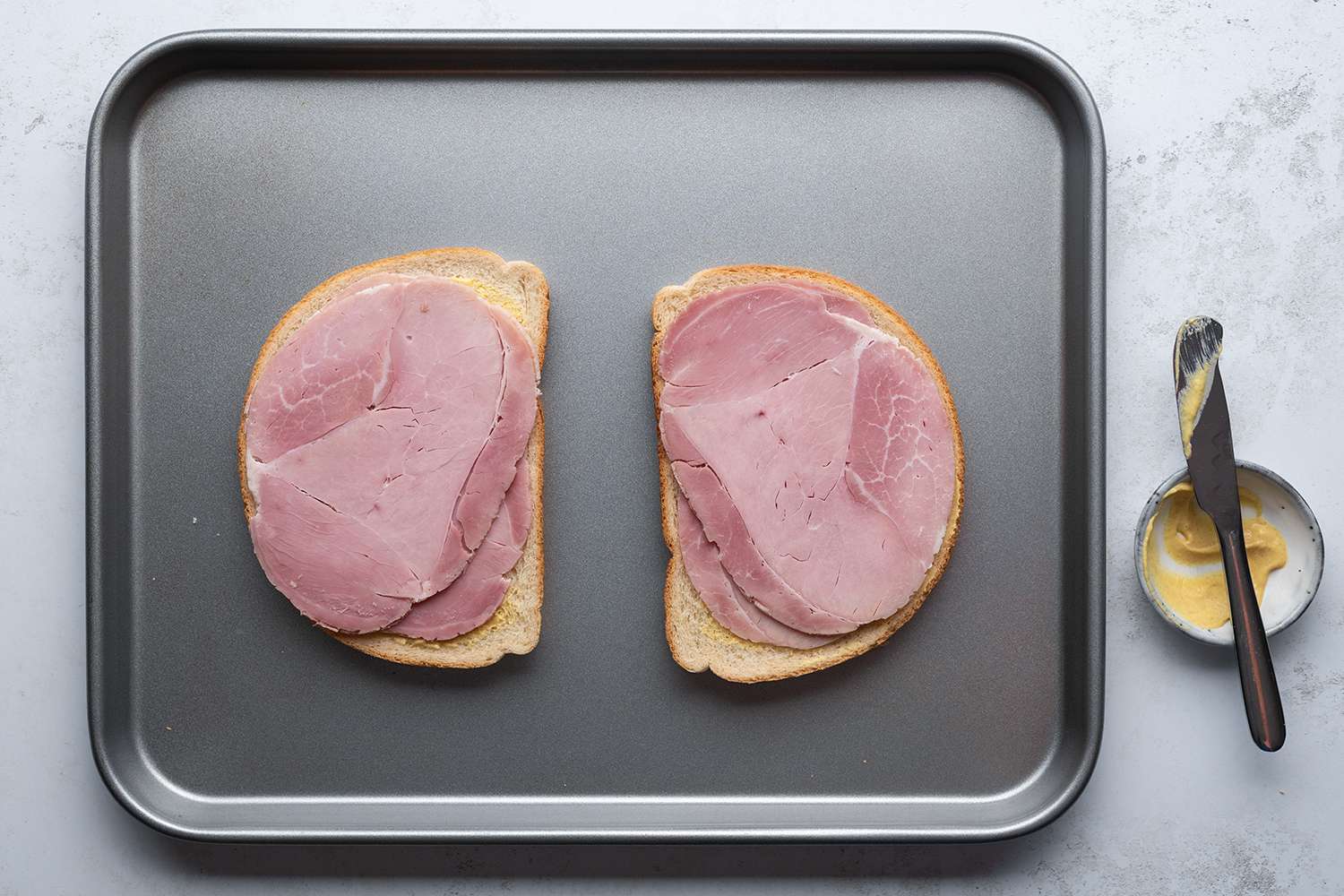 Bread slices with mustard and ham on a sheet pan