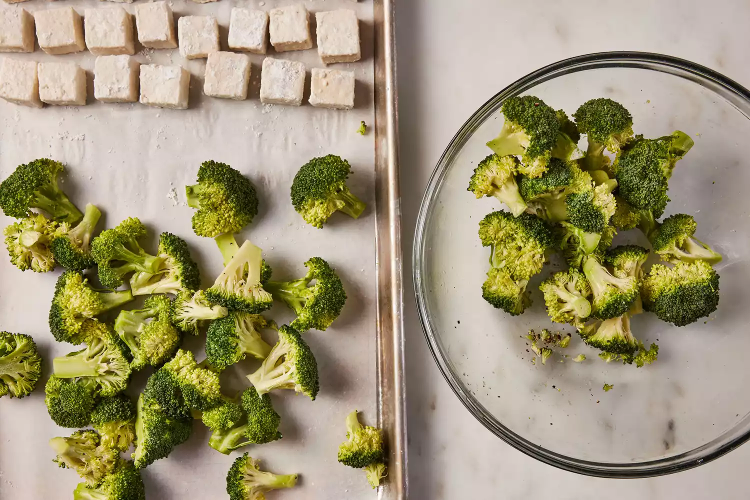 A bowl of broccoli florets next to a parchment paper-lined baking sheet with cornstarch-coated tofu and oil-tossed broccoli florets