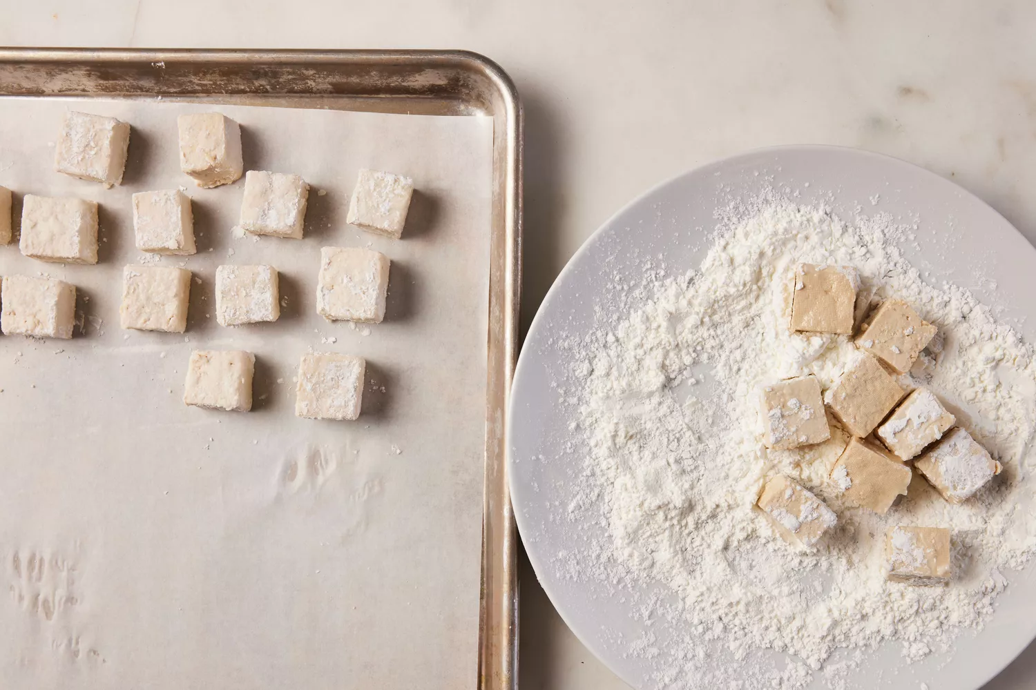 A large plate of cornstarch with cubed tofu next to a parchment paper-lined baking sheet with cornstarch coated tofu