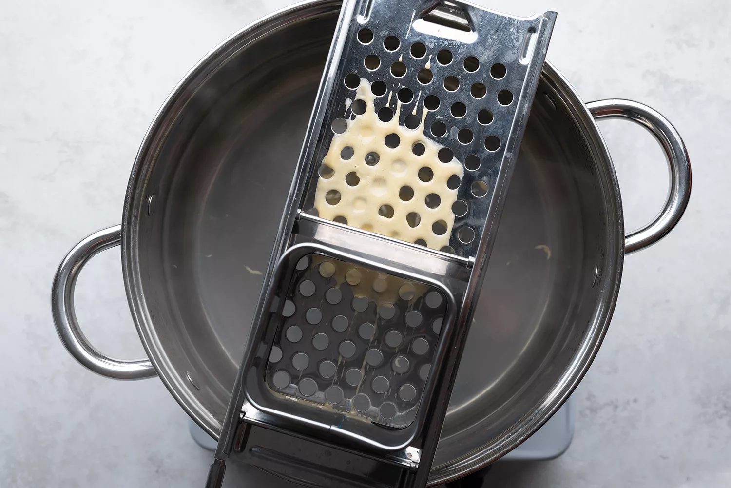 Massa do spaetzle sendo despejada em cima de um utensílio sobre uma panela com água no fogão