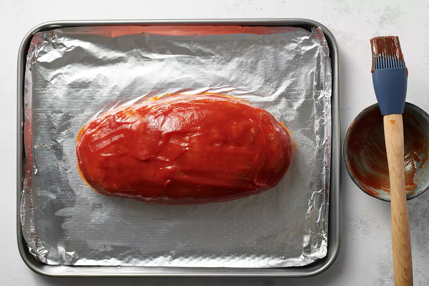 pão de carne cru coberto com ketchup em uma assadeira forrada de papel alumínio